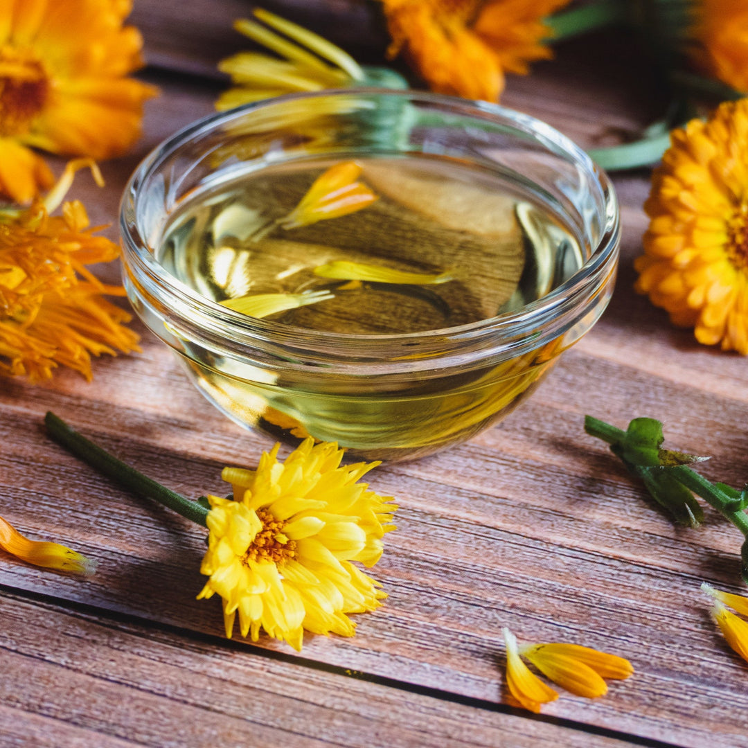 A clear glass bowl filled with yellow liquid, surrounded by fresh calendula flowers and petals on a wooden surface.
