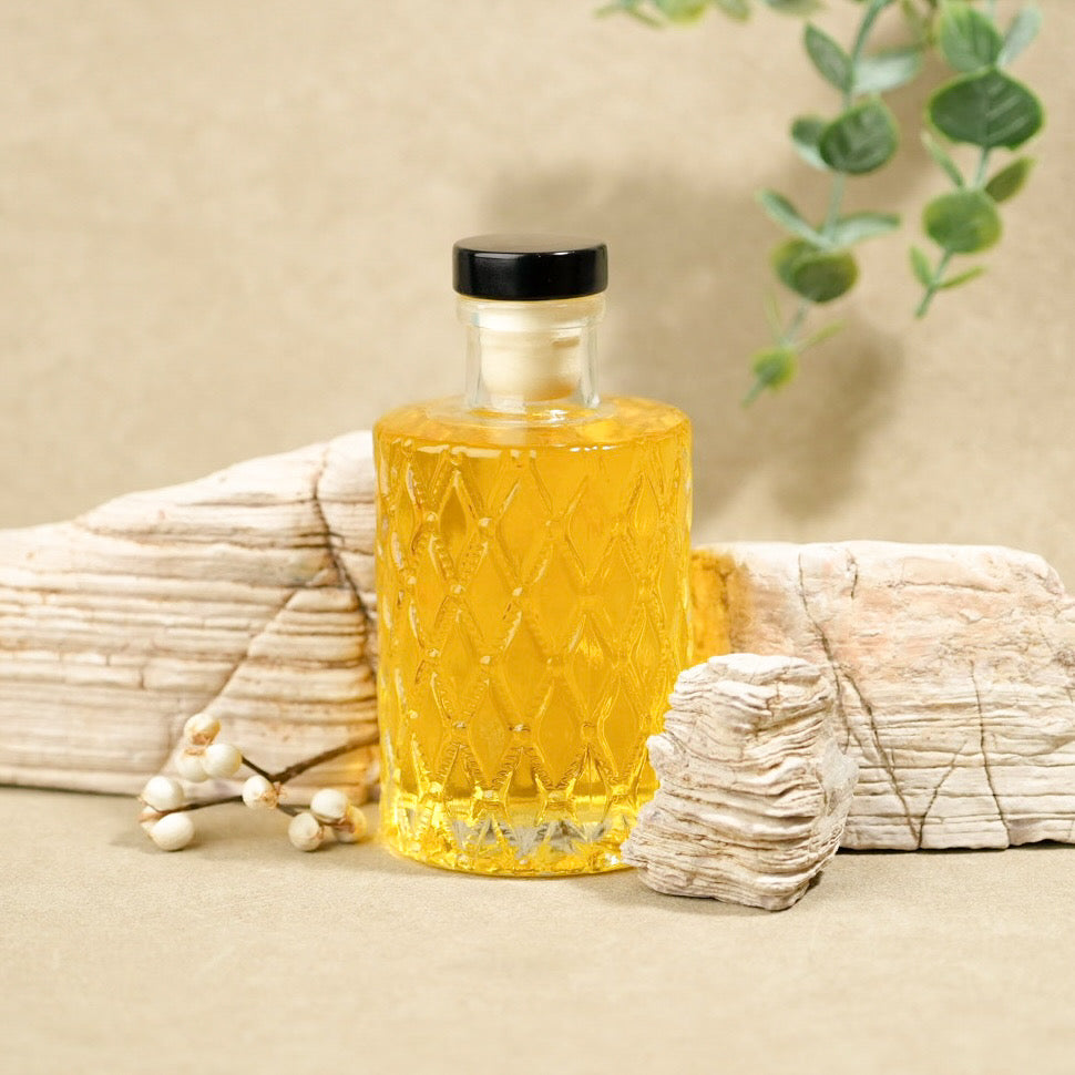 A clear glass bottle filled with golden yellow liquid, featuring a textured diamond pattern and a black cap, styled on a beige surface with driftwood, small white berries, and green leaves in the background.
