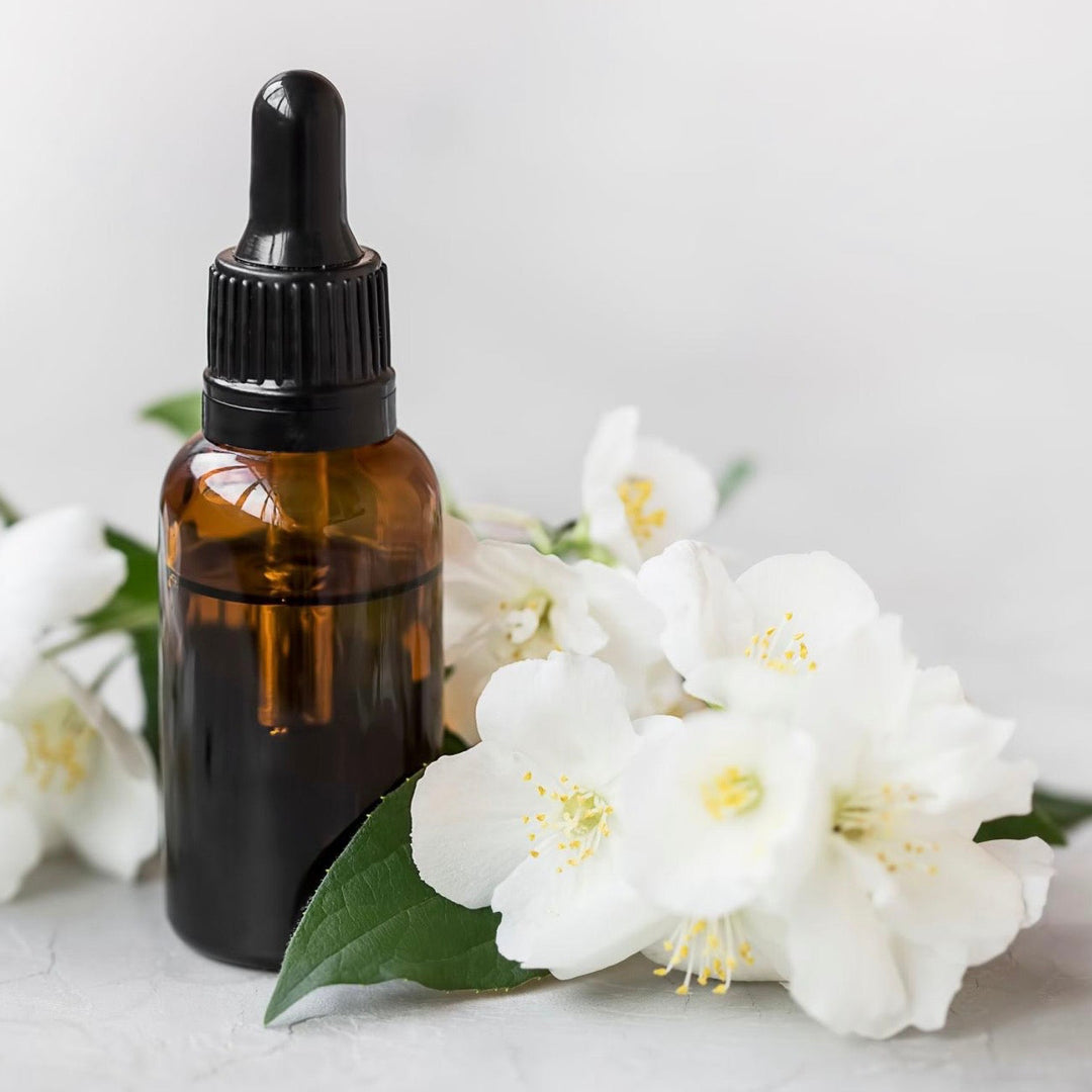 Brown glass dropper bottle with black cap surrounded by white flowers on a light gray background