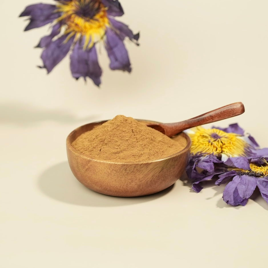 A small wooden bowl filled with fine brown powder, with a wooden spoon resting inside, set on a warm cream background with dried purple and yellow flowers around it.