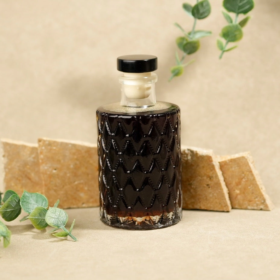 A clear glass bottle with a black screw cap and a dark brown liquid inside, featuring a textured chevron pattern on the front. The bottle is styled on a beige surface with eucalyptus leaves and crackers in the background.