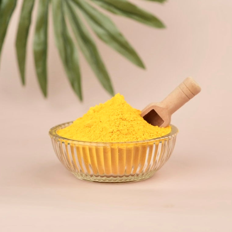 Glass bowl filled with yellow powder and a wooden scoop on a beige background with green leaves.