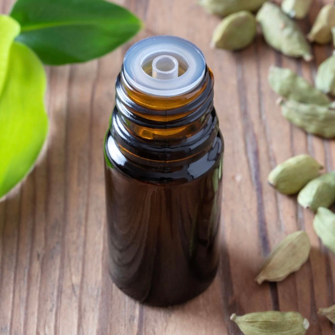 Brown glass roller bottle with a white cap on a wooden surface with cardamom pods and leaves.