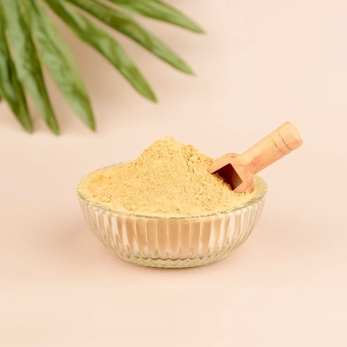 Glass bowl with beige powder and wooden scoop on a beige background with green leaves.