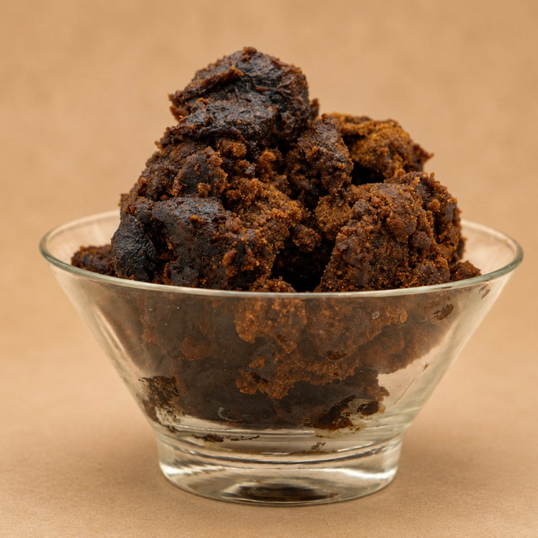 Dark brown textured chunks of kombo butter, piled in a small clear glass bowl against a warm beige background.