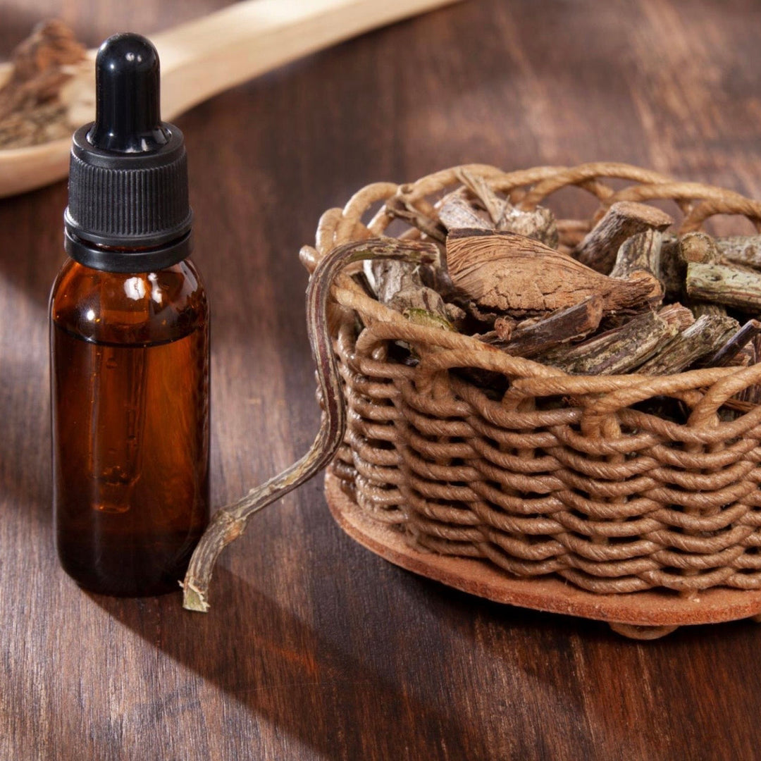 Brown glass bottle with dropper, wicker basket with dried herbs, and wooden spoon on a wooden surface.