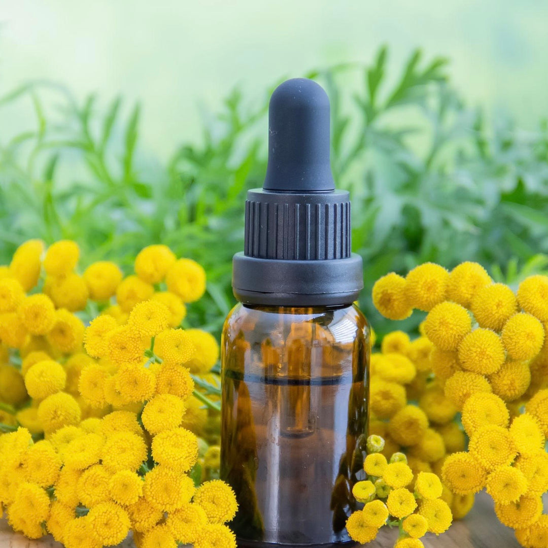 Brown glass dropper bottle with black cap surrounded by yellow flowers and green leaves.