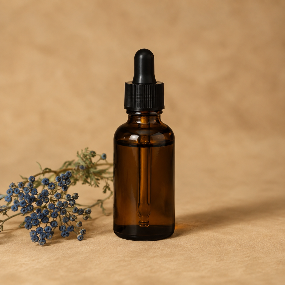 An amber glass dropper bottle with a black cap, standing on a textured beige surface next to a small sprig of dried blue flowers against a matching beige background.