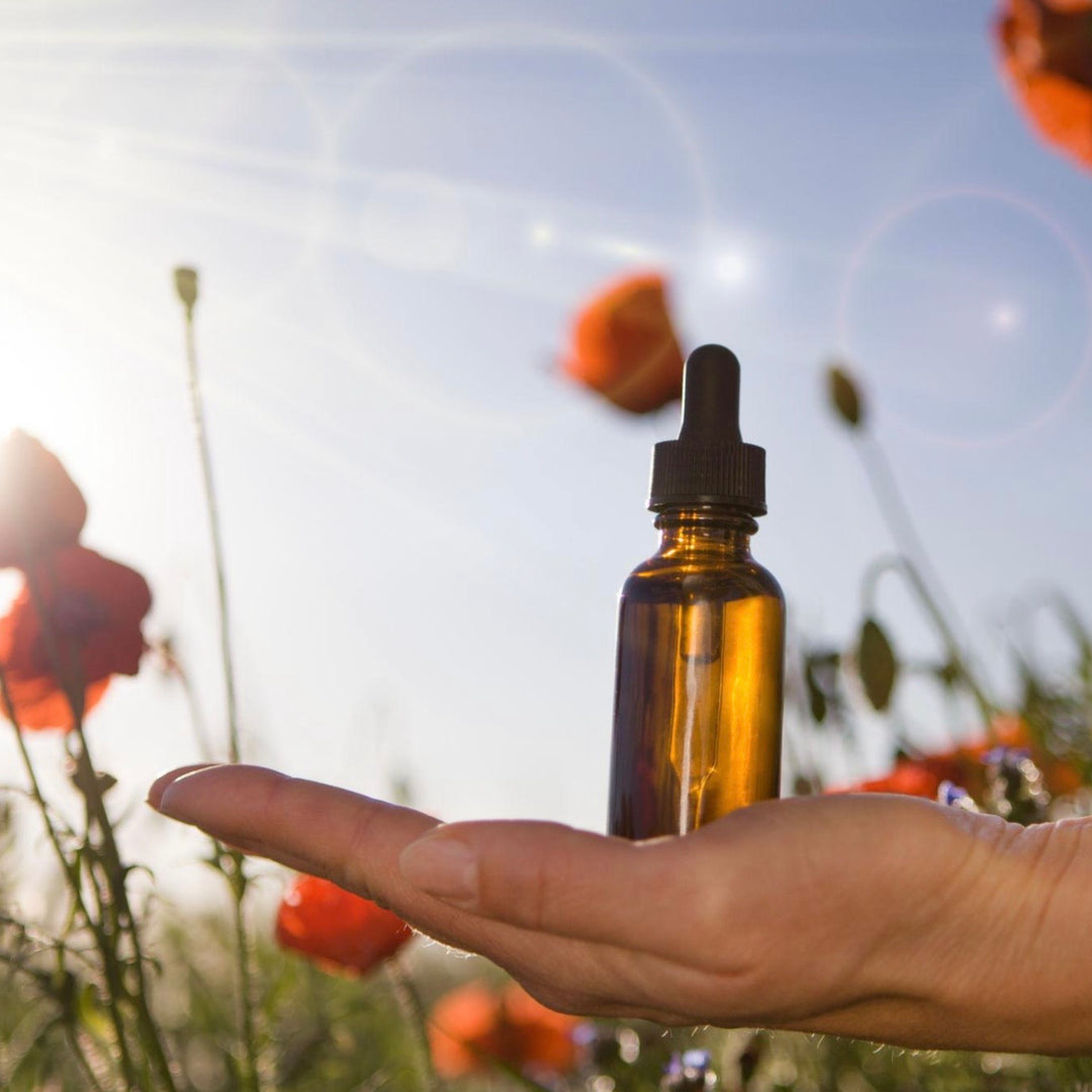 Hand holding a brown glass dropper bottle with flowers in the background