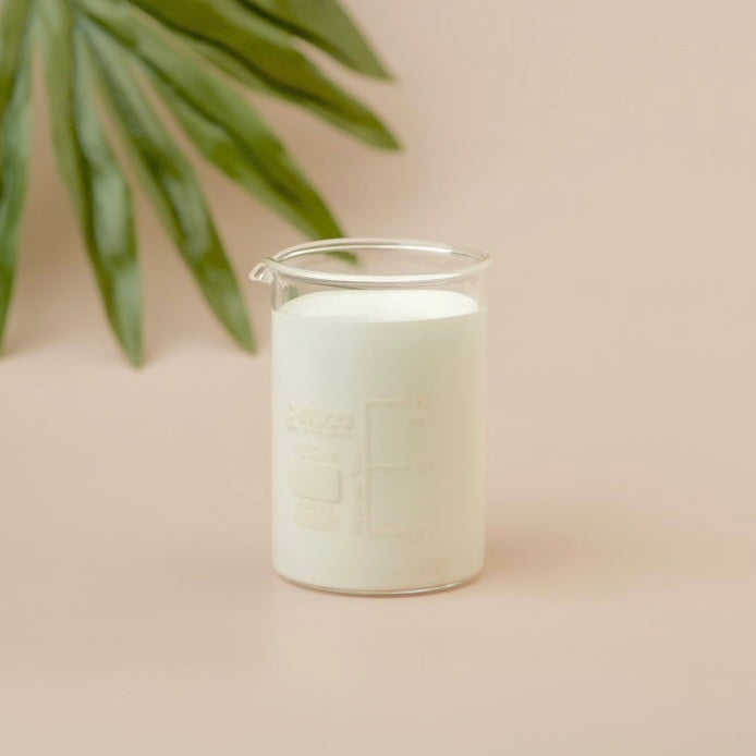White candle in a glass container on a beige surface with a blurred green plant in the background